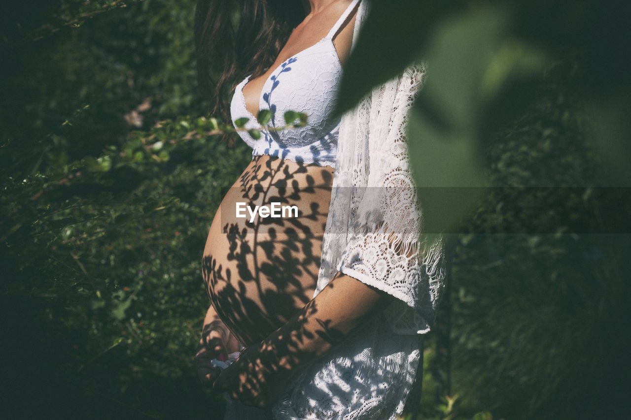 Midsection of pregnant woman standing amidst plants during sunny day