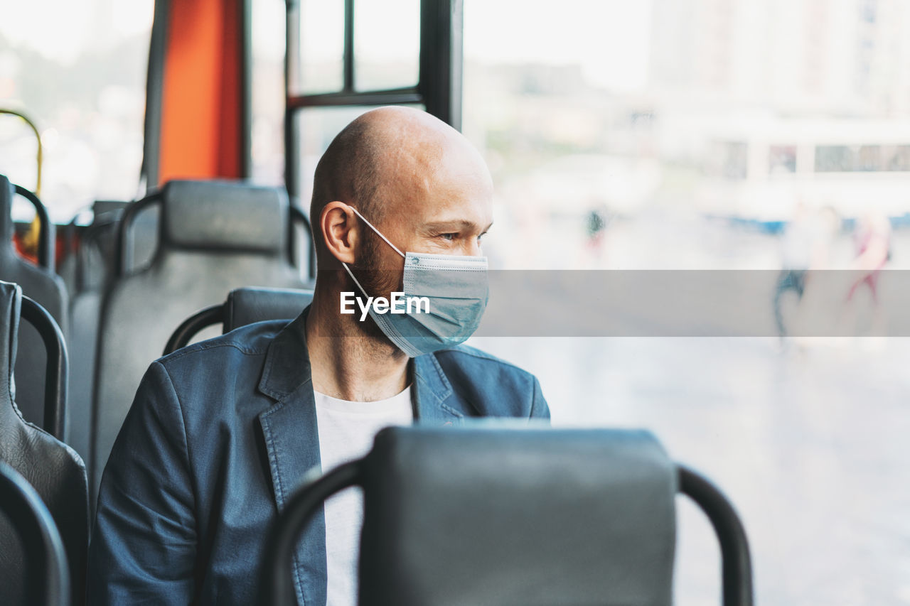 Adult bald bearded man in medical face mask looking out the window in bus