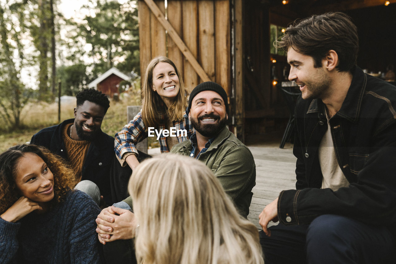 Smiling male talking to friends while sitting outside cottage during social gathering