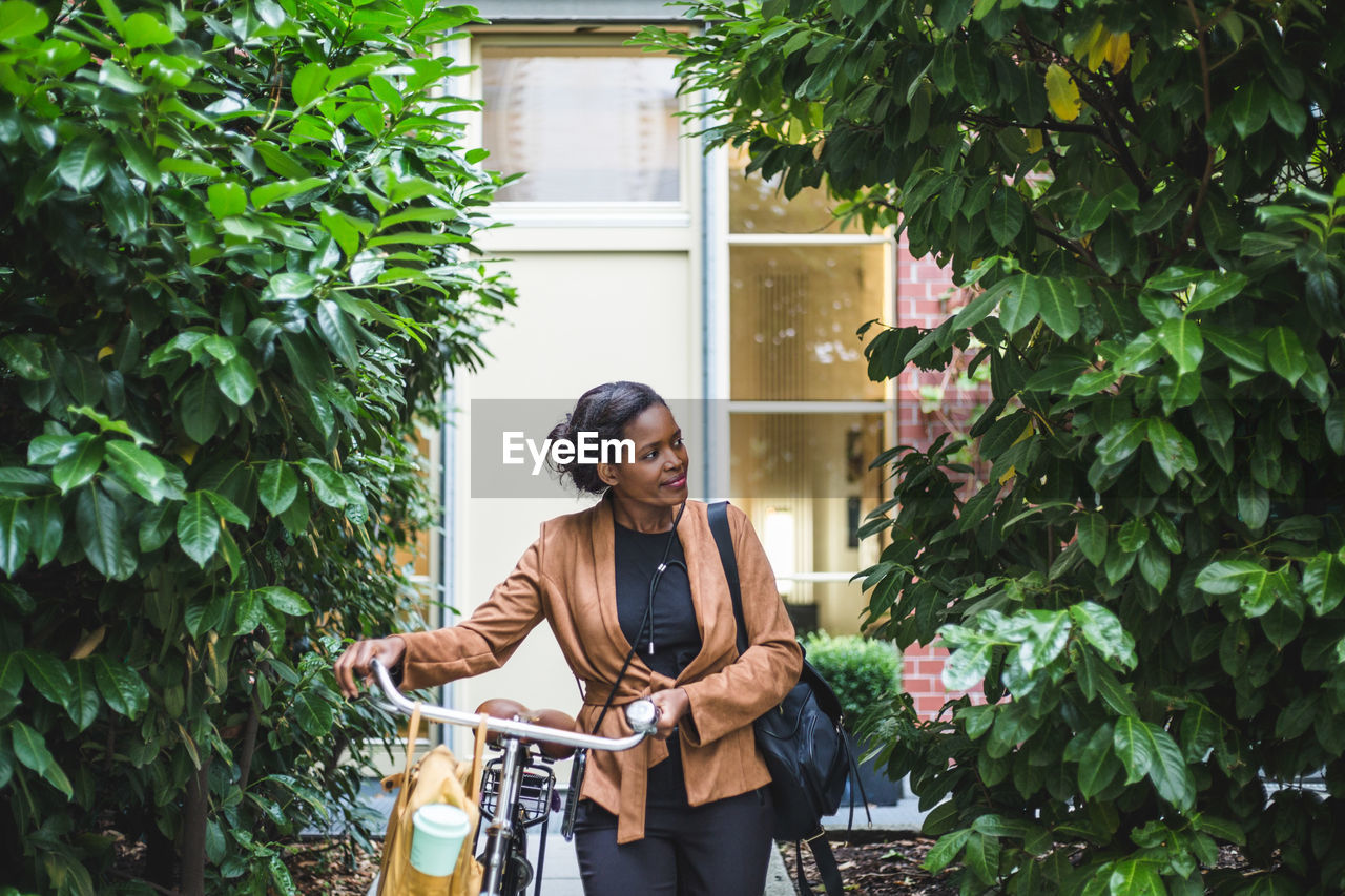 Smiling female architect looking away while standing with bicycle against house