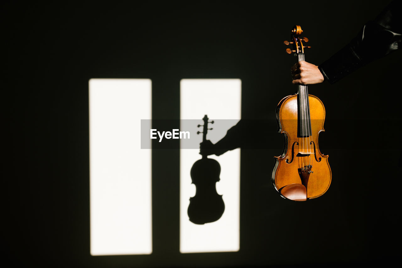 Crop unrecognizable musician in silk black shirt holding modern acoustic violin in outstretched hand in dark room against window on sunny day person