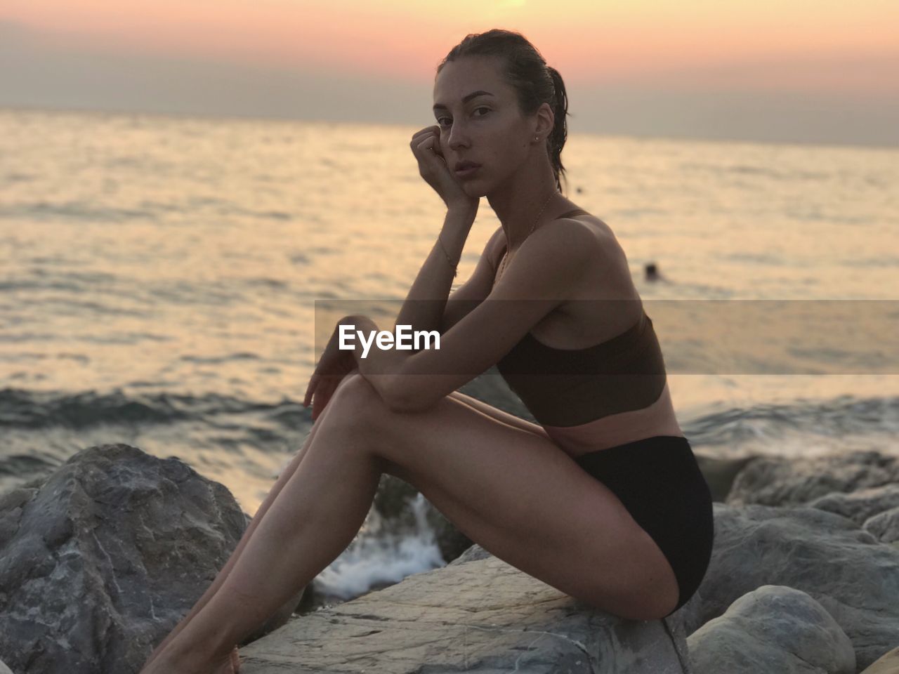 Side view portrait of young woman sitting on rock at beach during sunset