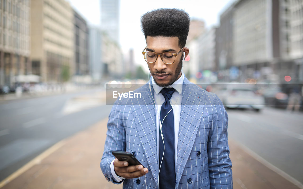 YOUNG MAN USING PHONE WHILE STANDING ON CITY STREET