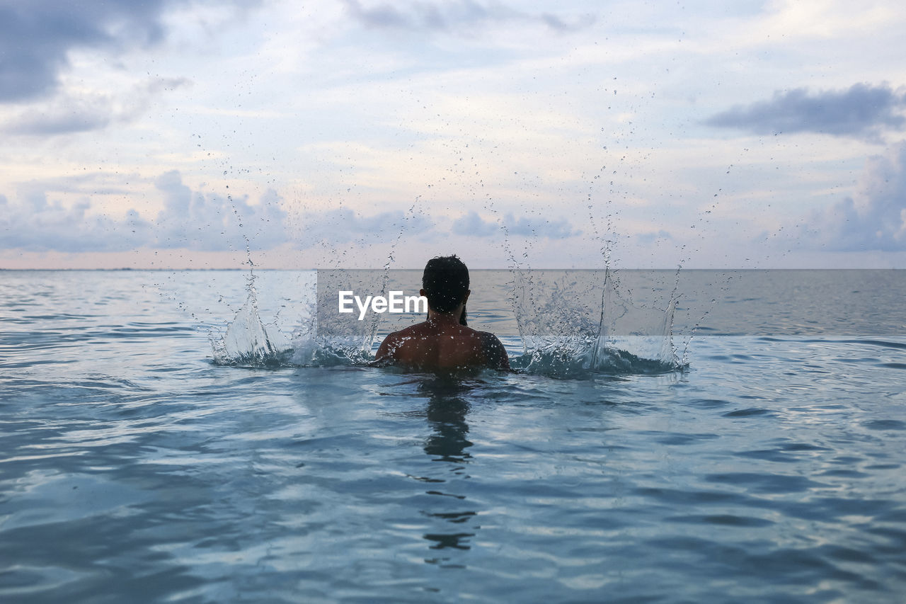 Rear view of shirtless man splashing water in sea against cloudy sky