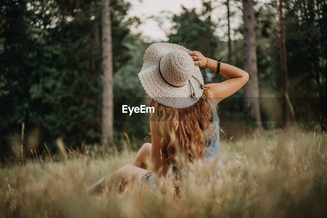 Rear view of woman wearing hat sitting on grassy field in forest