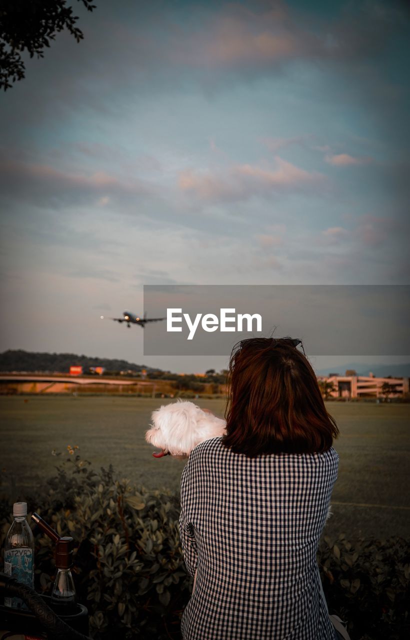 REAR VIEW OF WOMAN WITH AIRPLANE FLYING AGAINST SKY