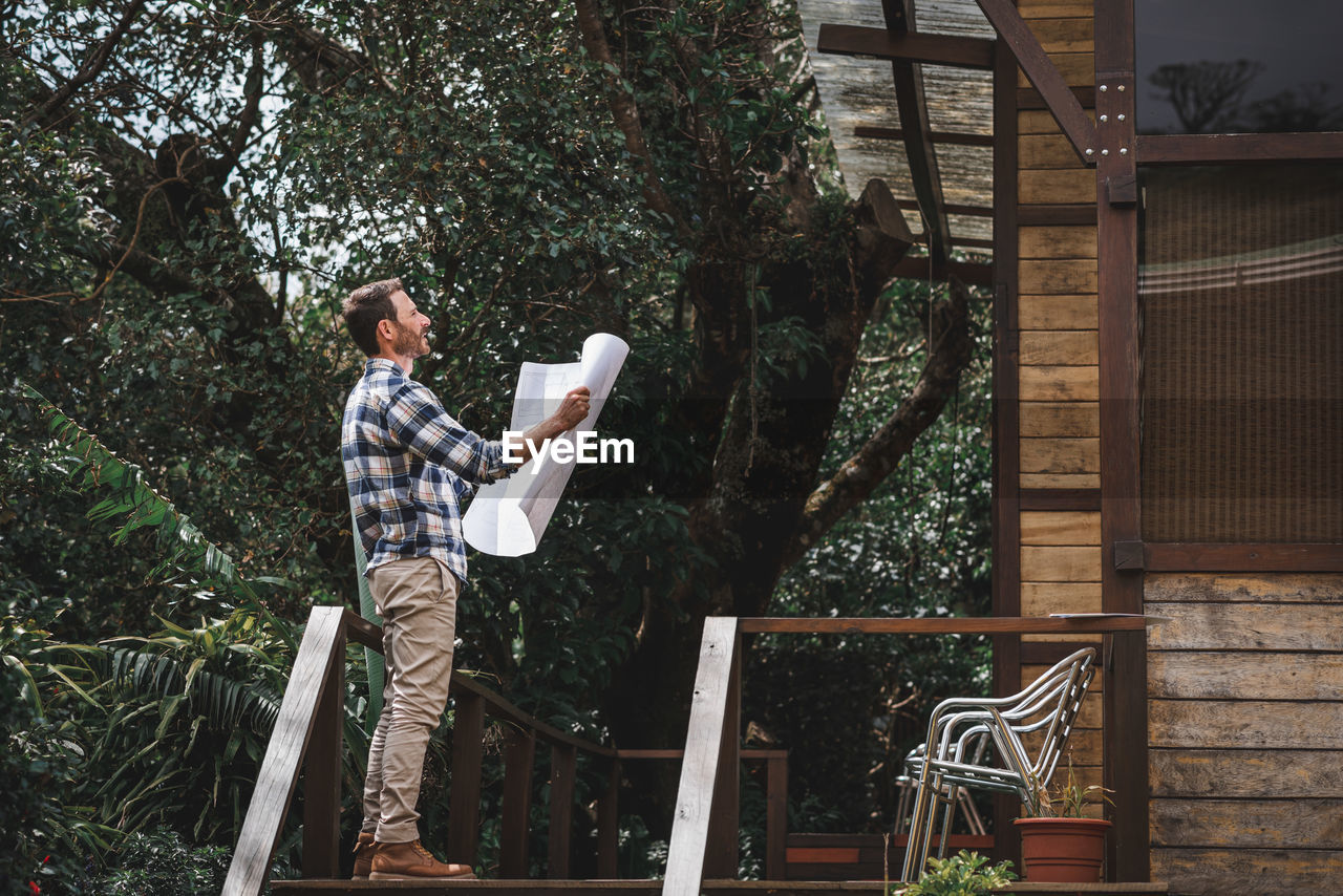 Low angle side view of male architect with paper draft standing on terrace of wooden house in forest