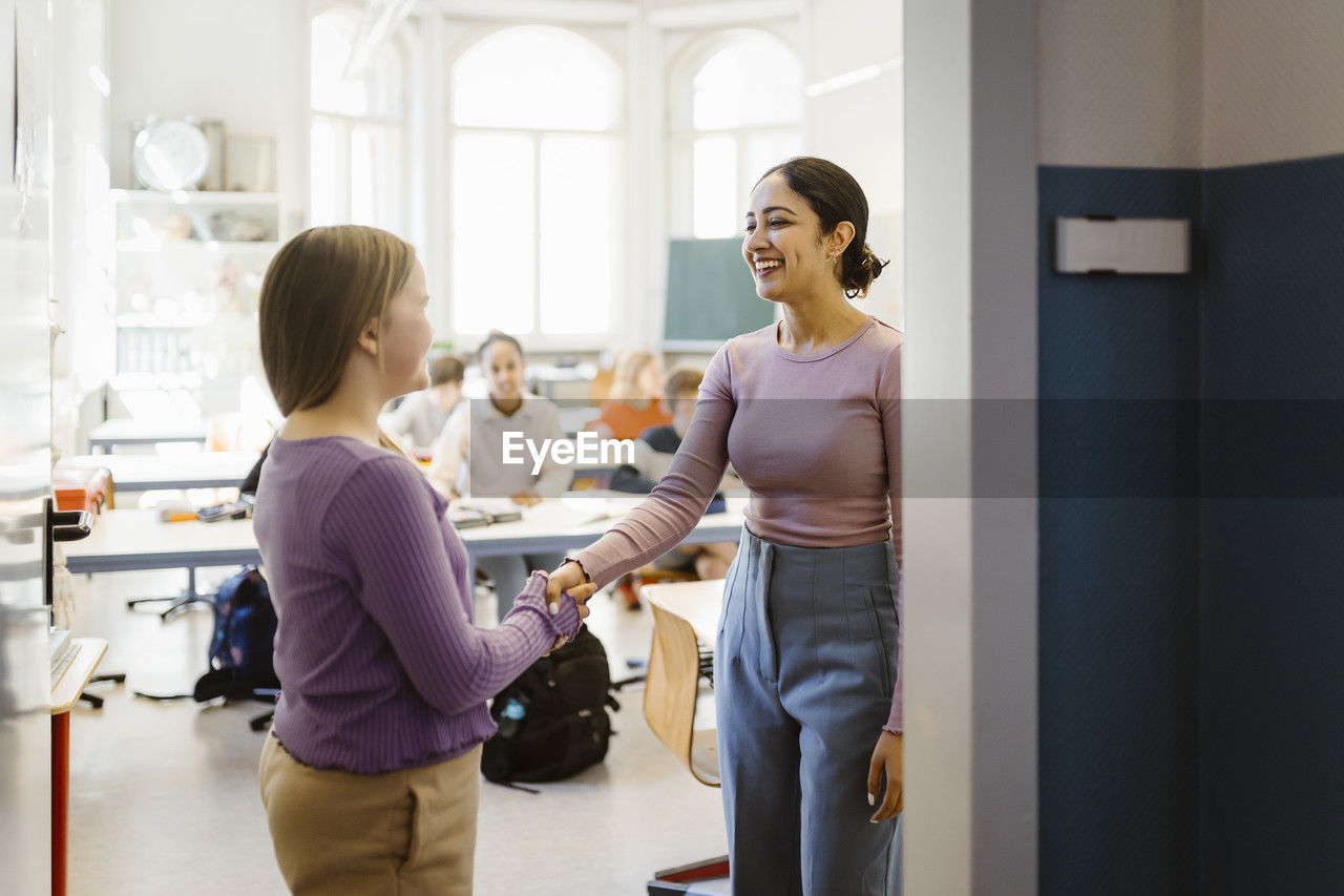 Smiling female teacher doing handshake with student standing in classroom