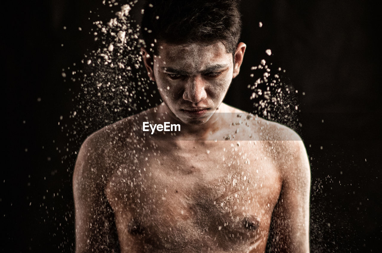 Close-up of shirtless man with flour on body standing against black background