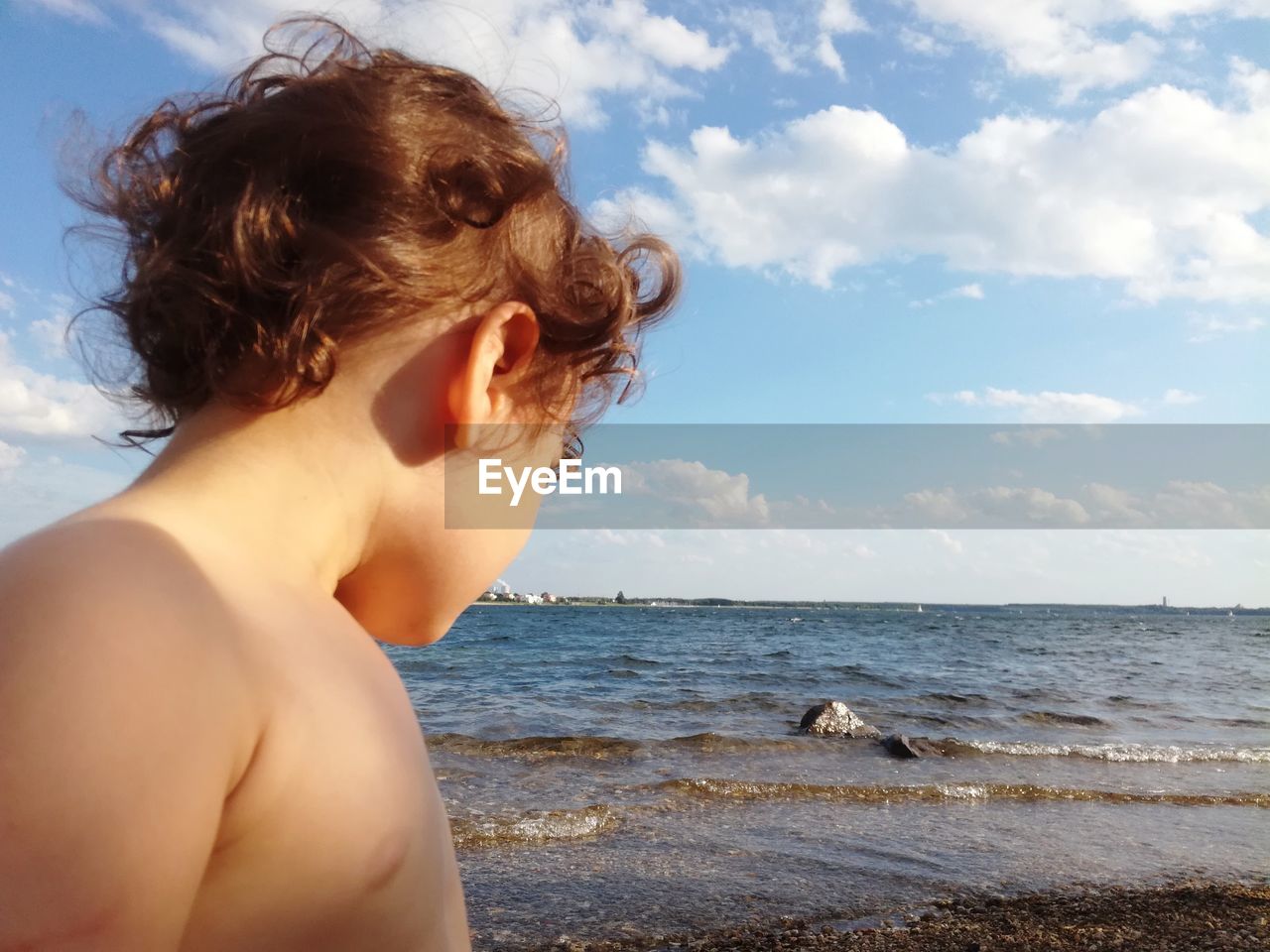 Side view of shirtless boy standing at beach against sky