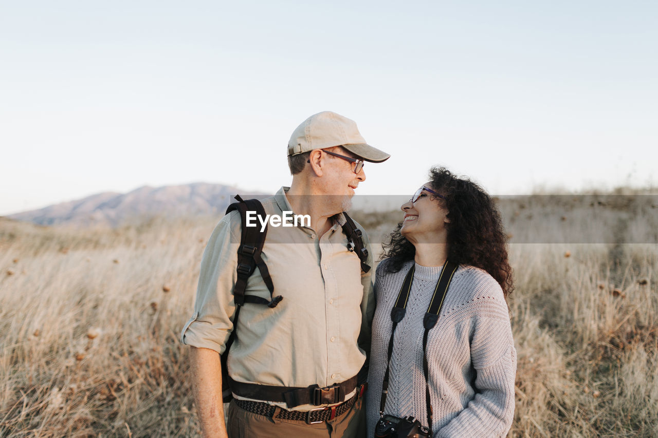 Cheerful mature couple in casual clothes embracing and standing on grassy meadow in countryside