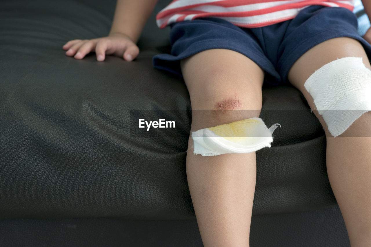 Midsection of boy showing wound while sitting on sofa with bandage on knees