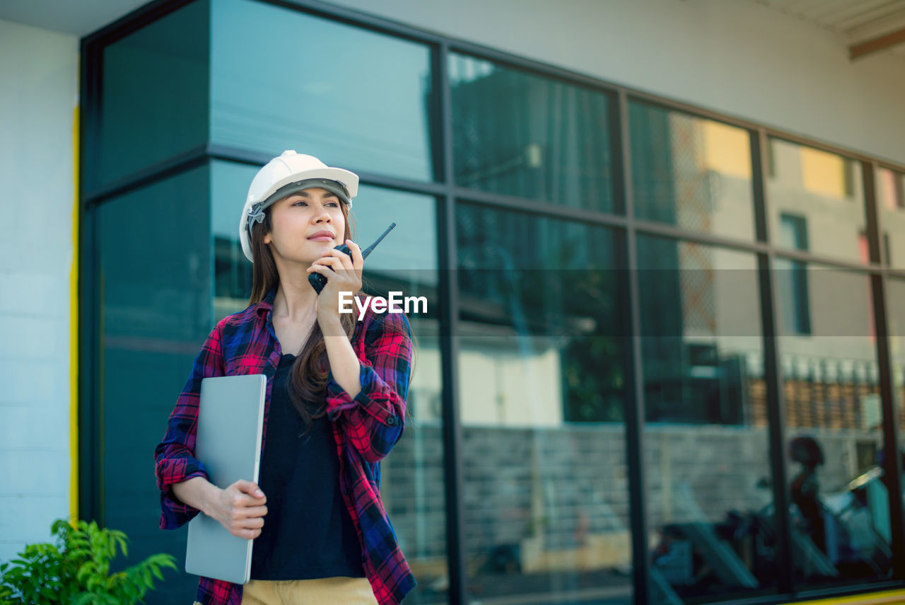 YOUNG WOMAN LOOKING AT CAMERA WHILE STANDING AGAINST BUILDINGS