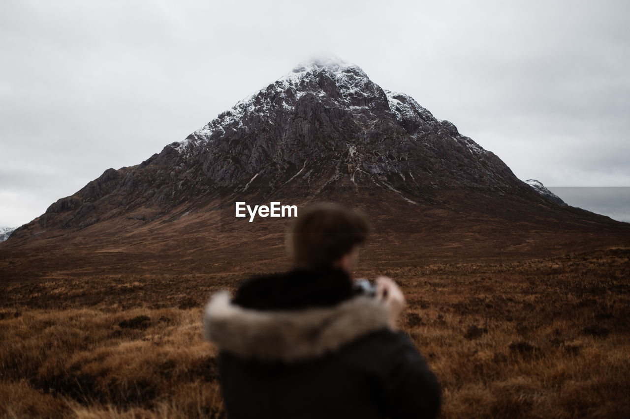 Back view of unrecognizable blurred tourist in warm clothes admiring landscape of mountain with snowy peak in scottish highlands in fall