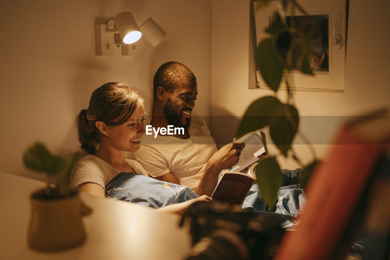 Smiling couple reading books by illuminated sconce mounted on wall at home