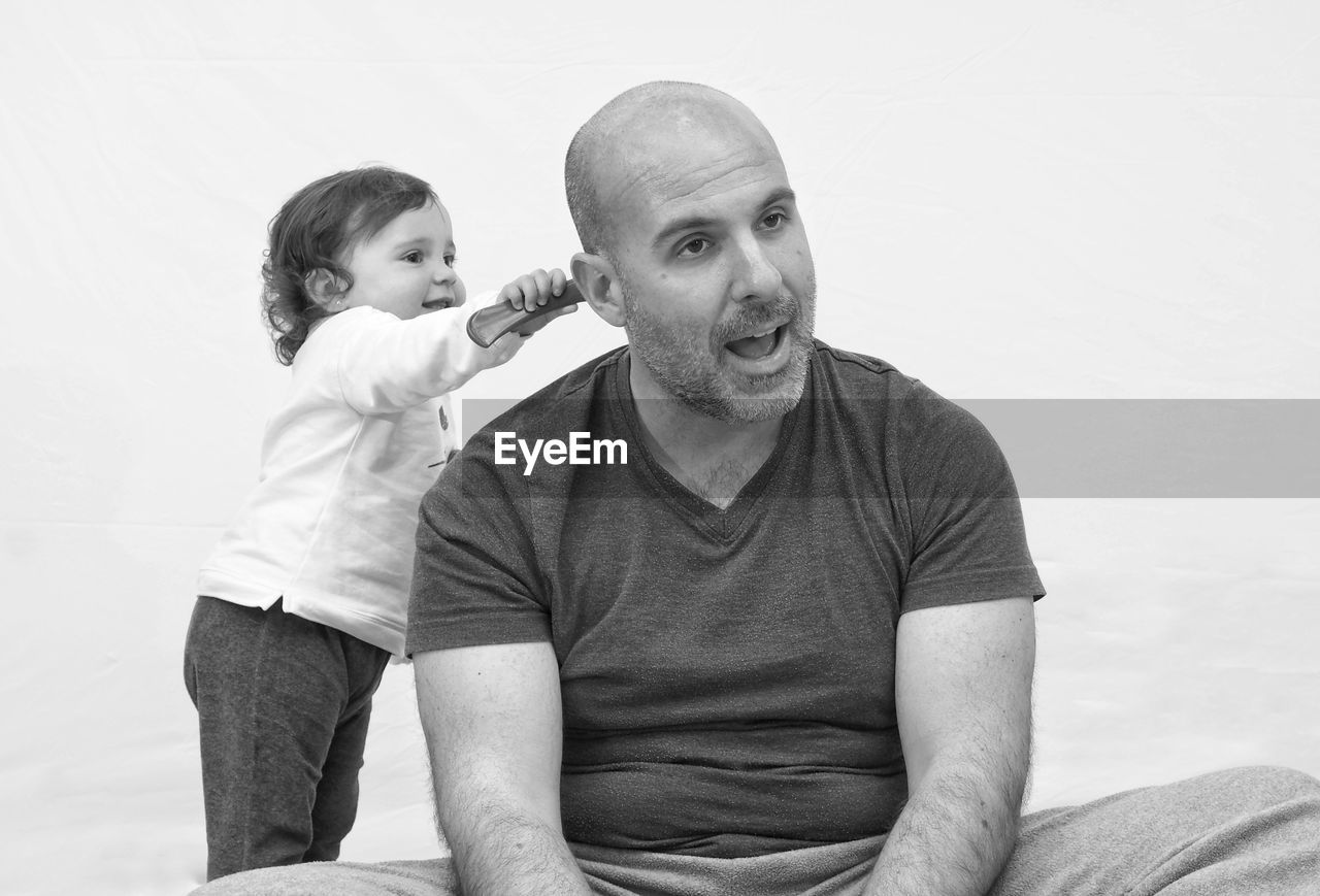 Cute girl combing hair of father while sitting against wall at home