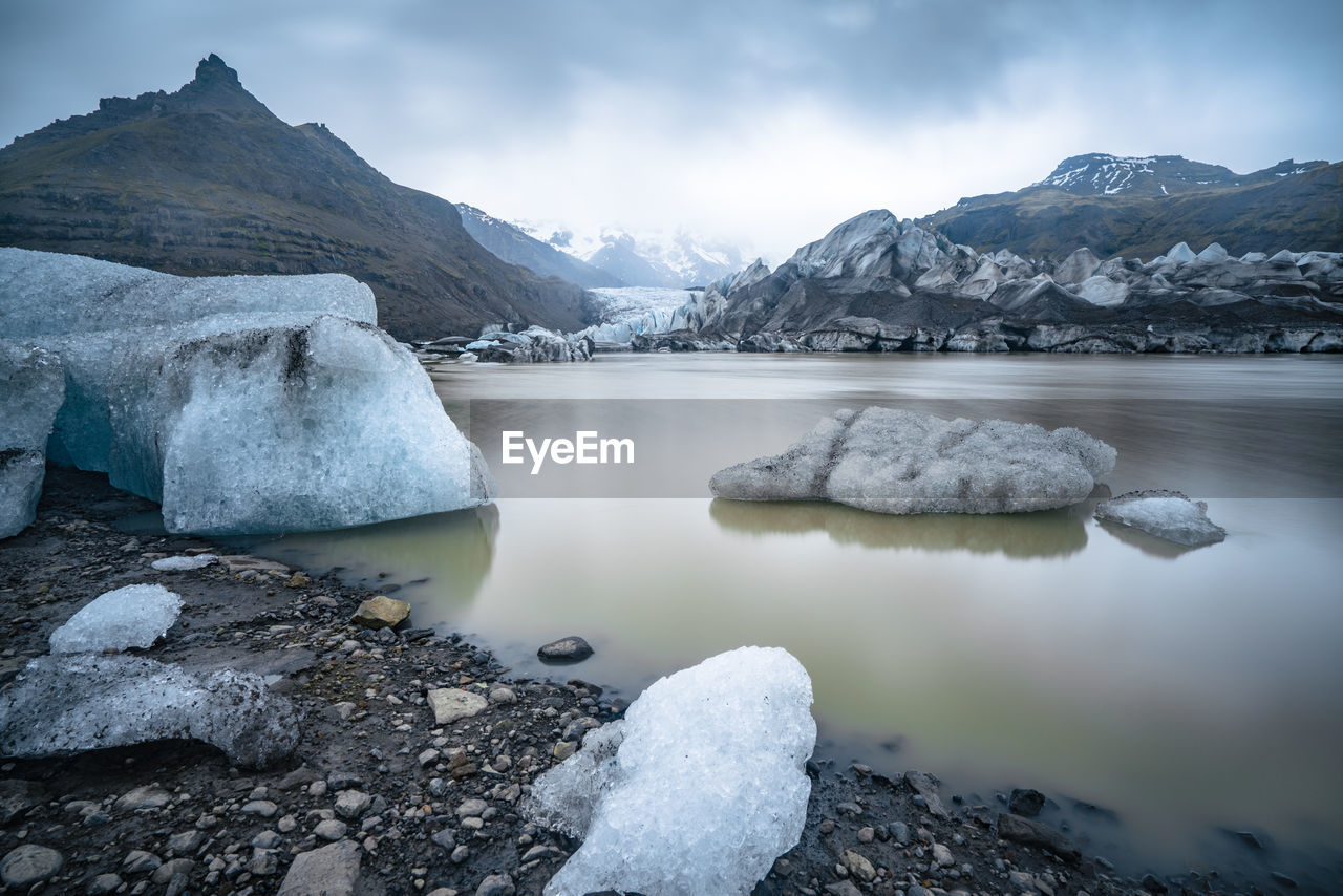 Ice floating in muddy waters of glacier lake with glacier in background on a cloudy and rainy day.