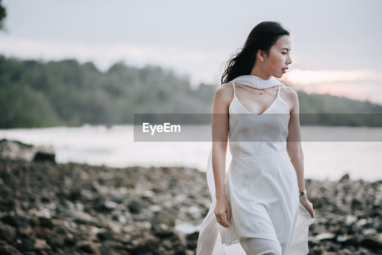 Young woman walking on pebbles at beach against sky during sunset