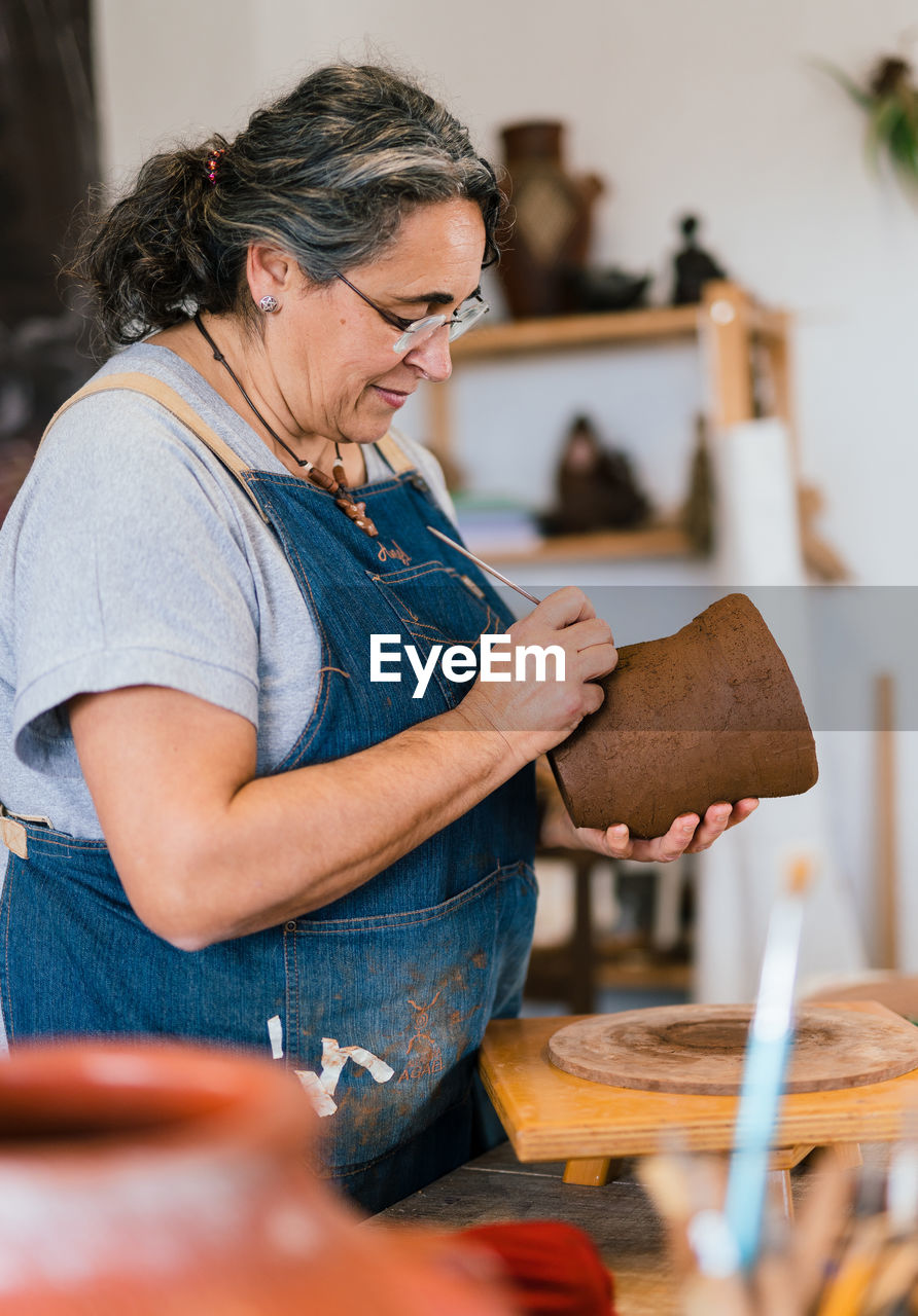 Mature female craftswoman ceramist in apron using tool and creating pattern on clay pot in workshop