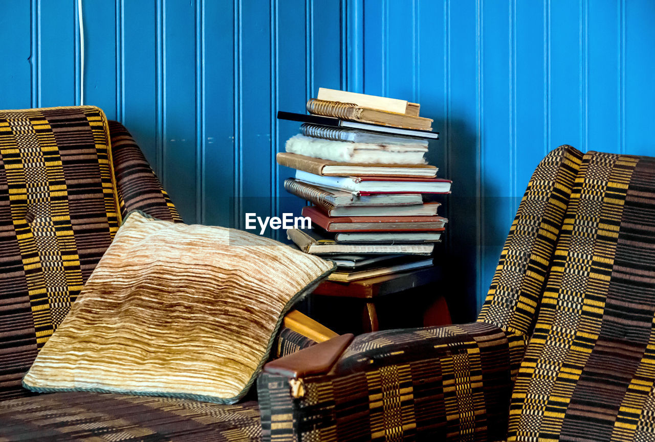 Two  armchairs stand in the corner of a blue-panelled room, a stack of books lies on a corner table.