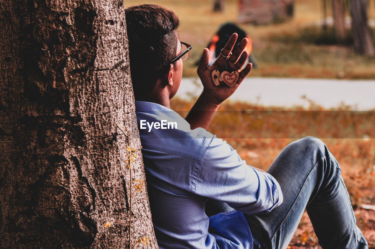 Rear view of young man sitting against tree trunk while looking at key ring