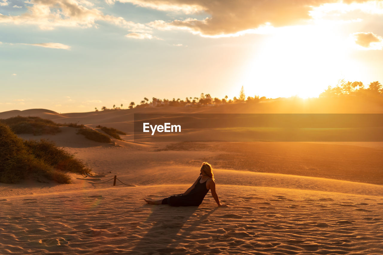 Full length of woman sitting on sand against sky during sunset