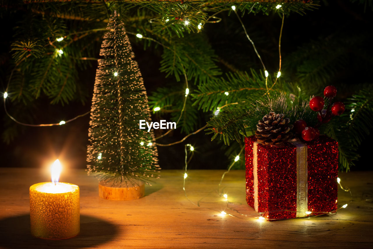 A red christmas box with a gift, a burning candle on background of a fir tree with christmas lights