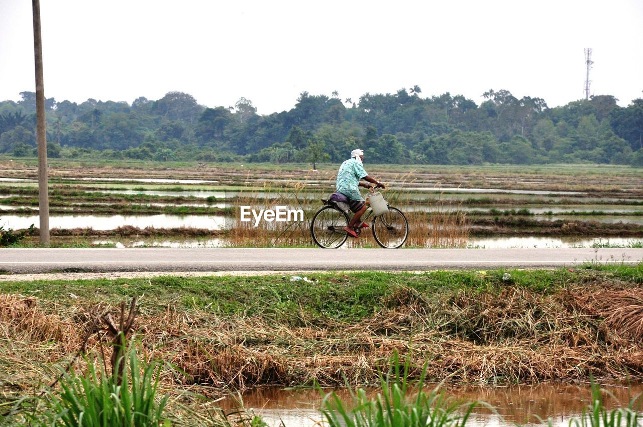 Man riding bicycle on road amidst field against clear sky