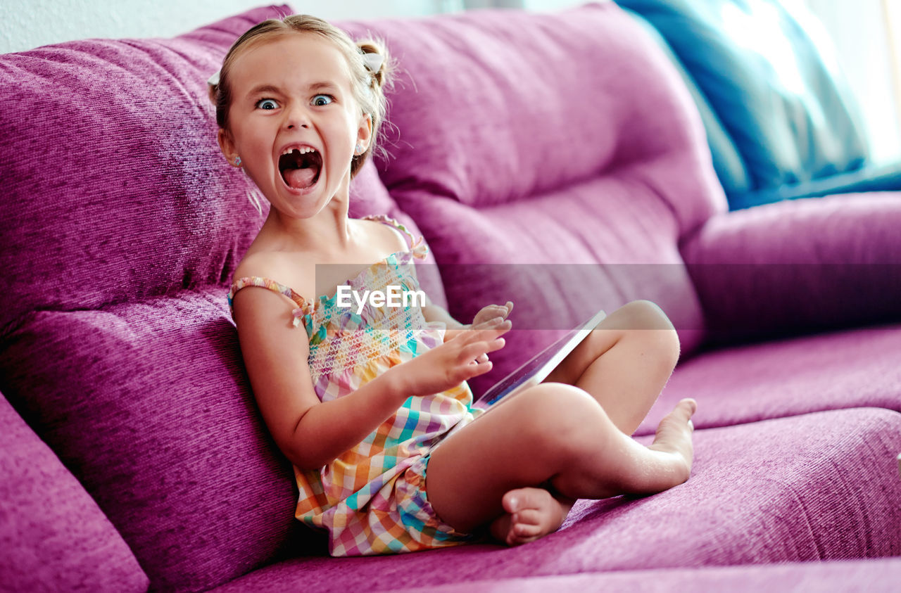 Portrait of cute girl using digital tablet sitting on pink sofa with mouth open at home