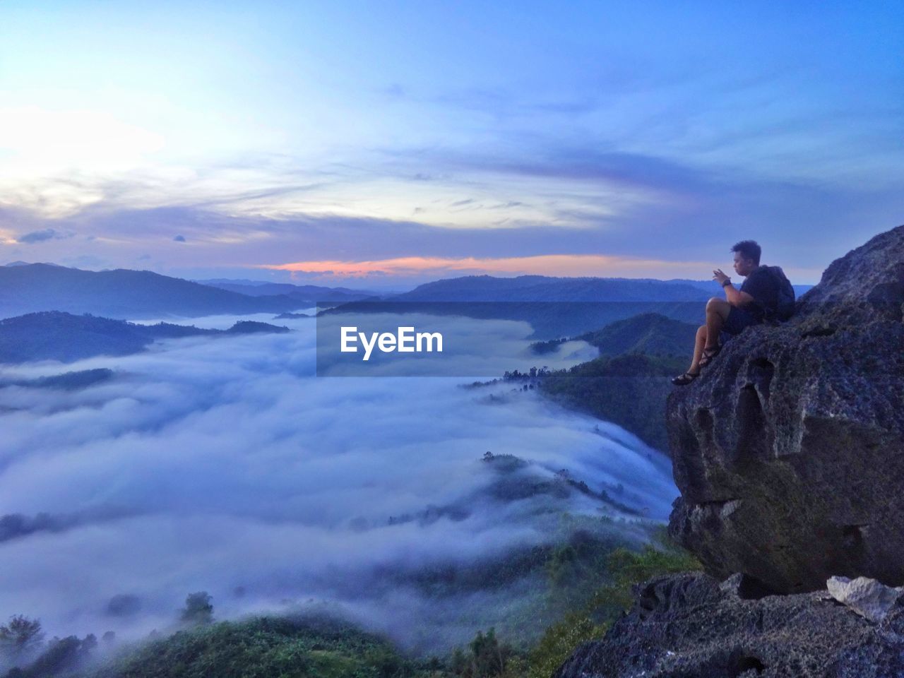 Side view of man looking at cloudscape while sitting on mountain during sunset