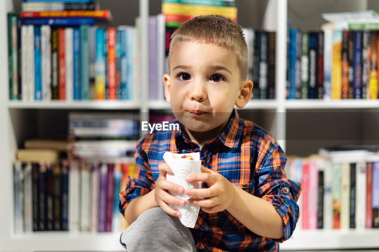 Portrait of boy eating snack against bookshelf