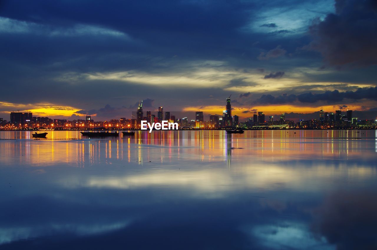 Illuminated buildings by sea against sky at sunset