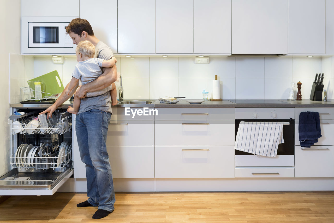 Full length of father working in kitchen while carrying baby at home