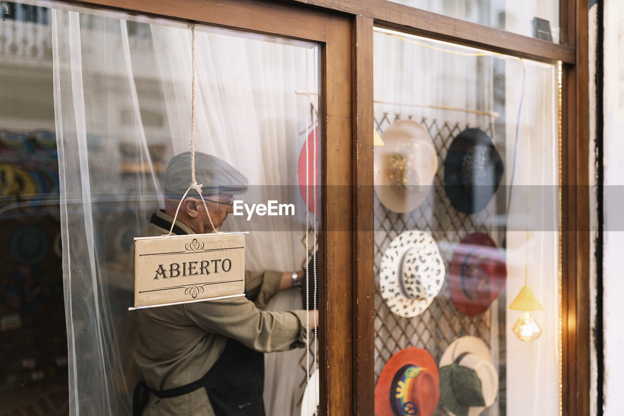 Through glass side view of concentrated mature male seller hanging stylish assorted hats behind showcase in fashion boutique