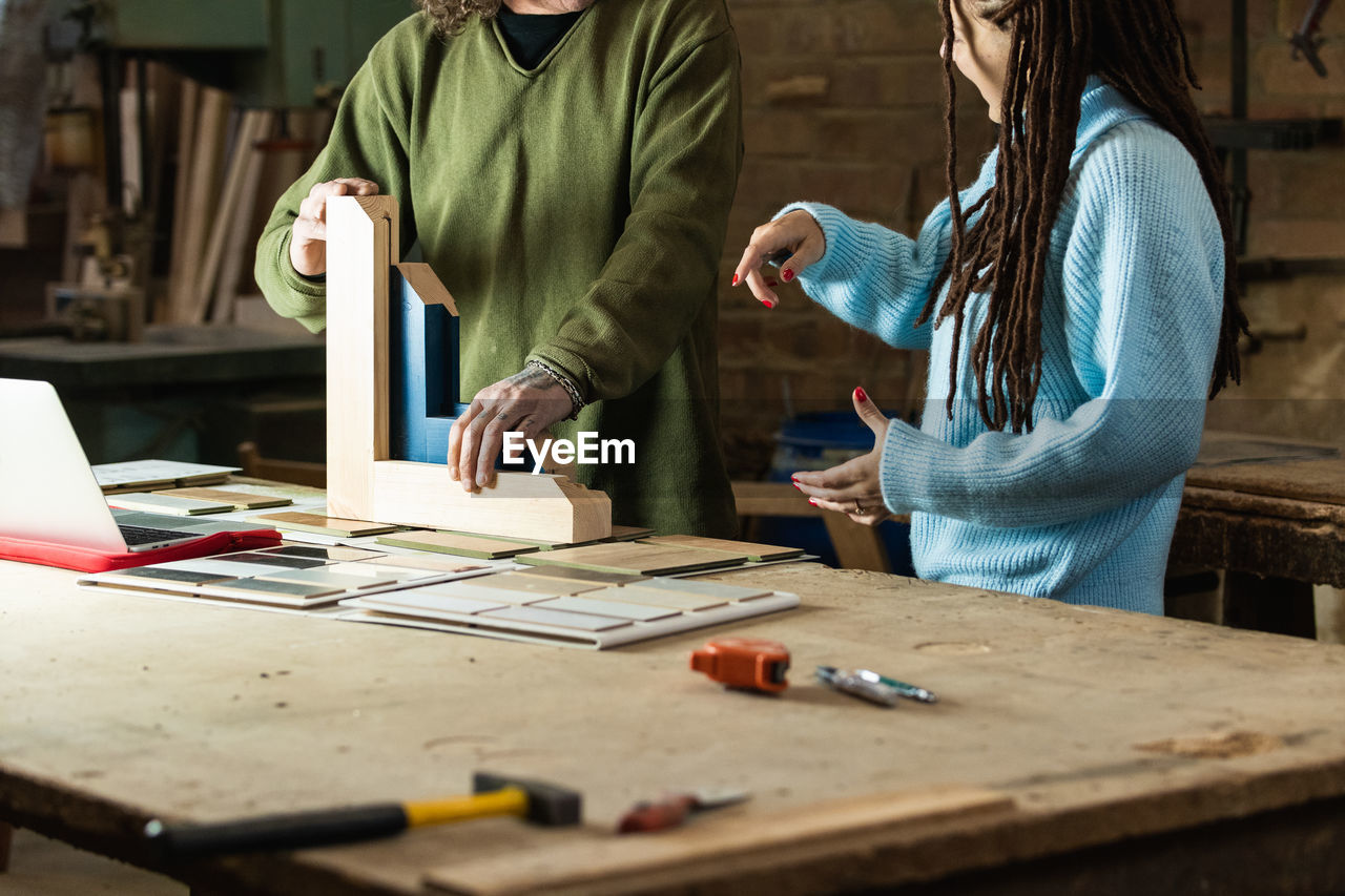 Crop carpenter standing at workbench with wooden detail and tools with laptop while discussing order with customer