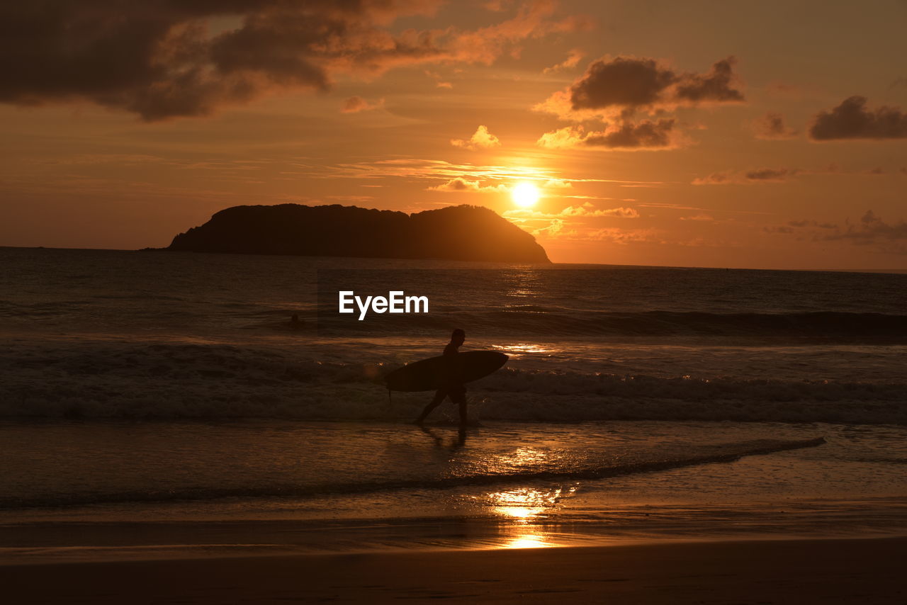 Silhouette man with surfboard walking at beach against sky during sunset