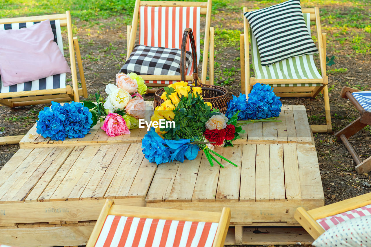 Beautiful artificial bouquets of various colors placed on wooden table among deck chairs in garden. 