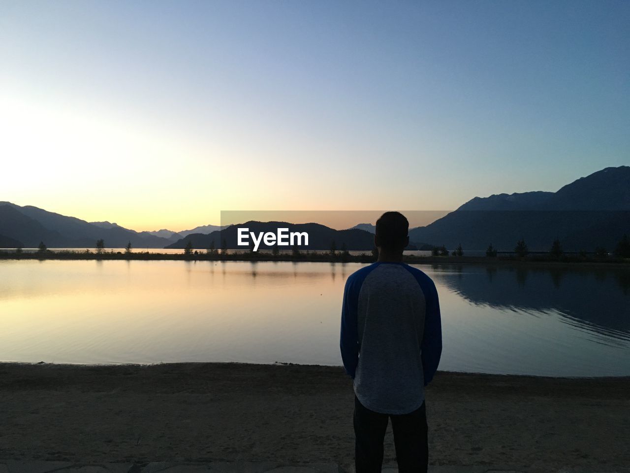 Rear view of man looking at lake against clear sky during sunset