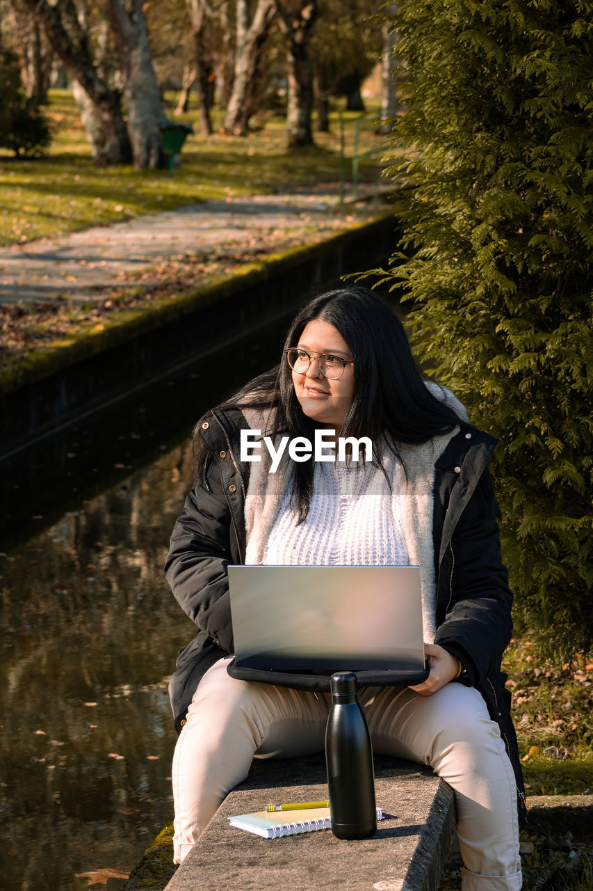 YOUNG WOMAN USING PHONE WHILE SITTING ON TREE