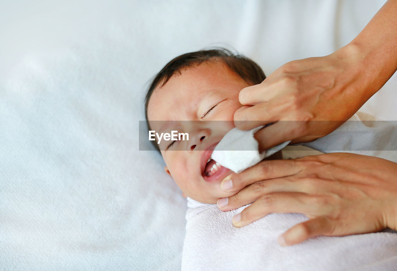 Cropped hand of mother cleaning baby boy teeth lying on bed at home