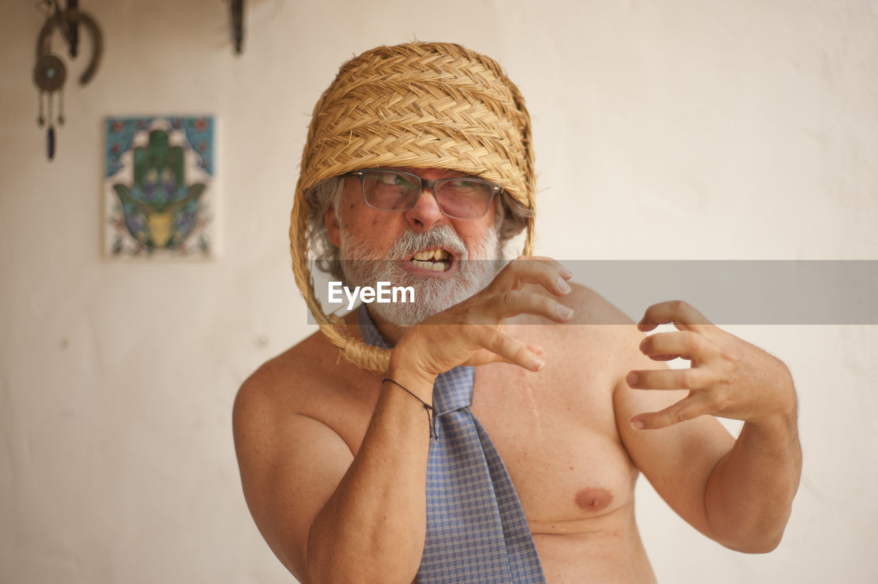 Angry shirtless man wearing basket on head while gesturing against wall
