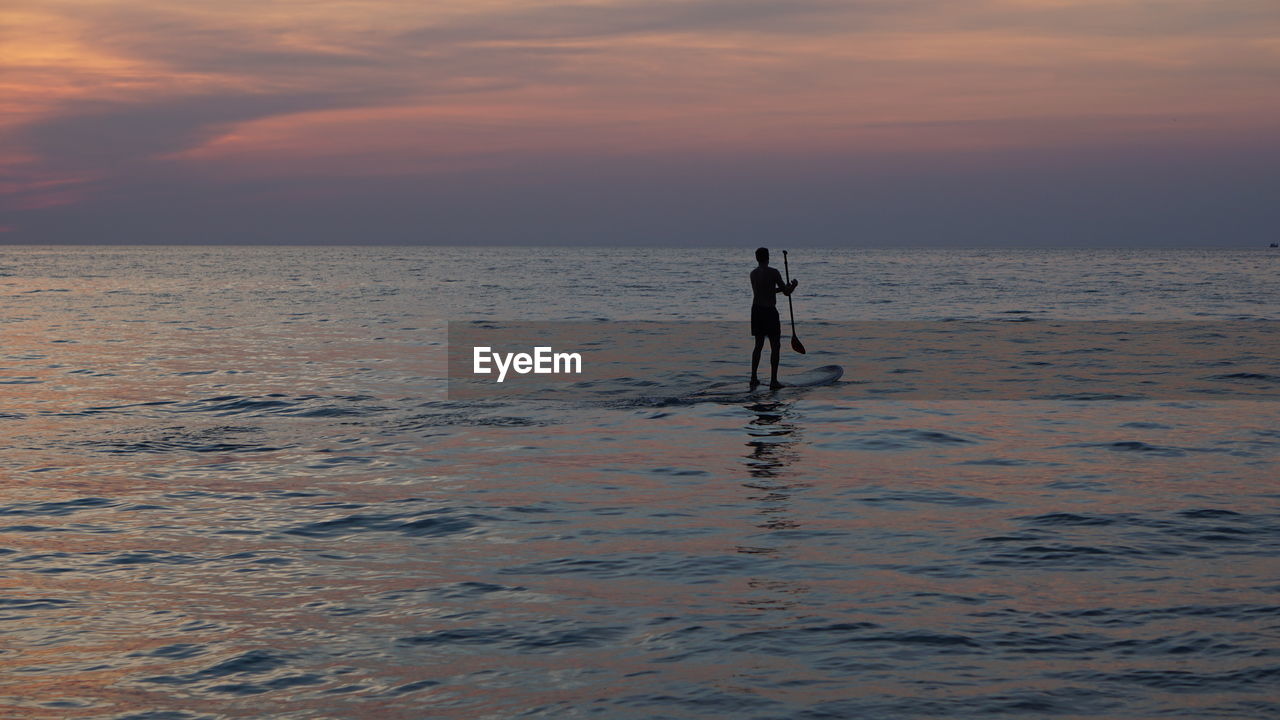 SILHOUETTE MAN STANDING IN SEA AGAINST SKY AT SUNSET