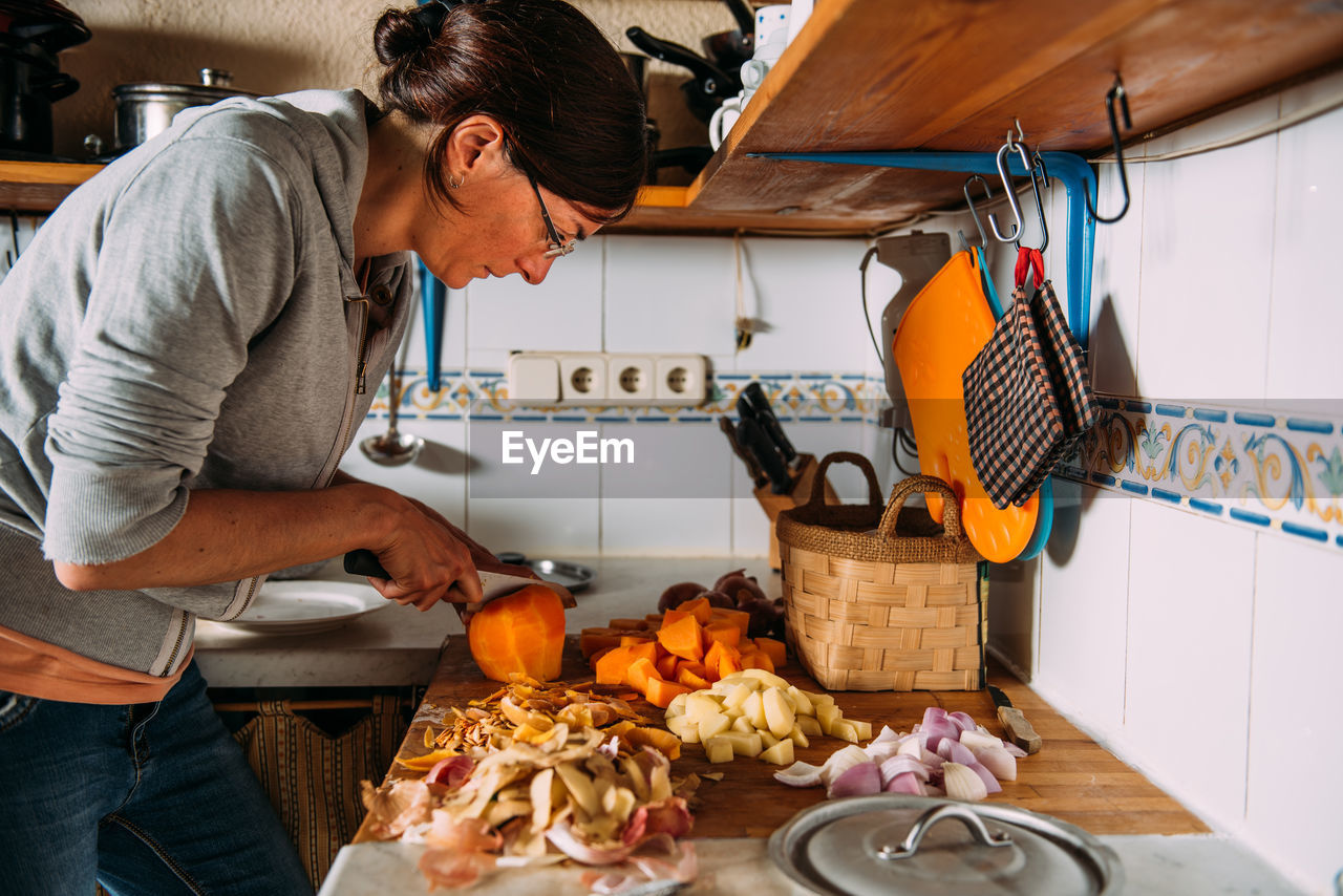 Side view of adult housewife chopping fresh pumpkin on table with various vegetables while preparing healthy natural cream soup at home