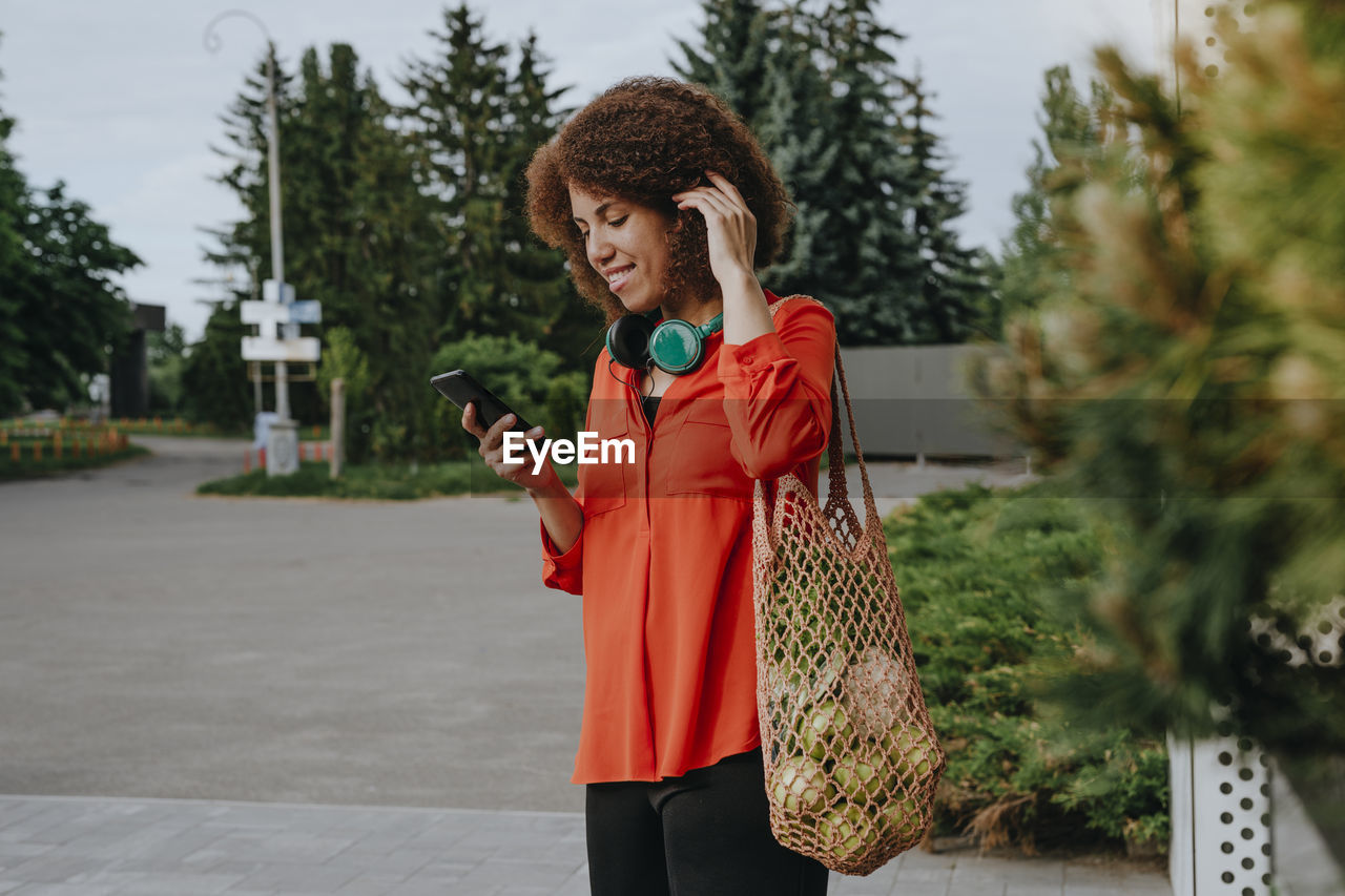 Young woman carrying apples in mesh bag and using mobile phone