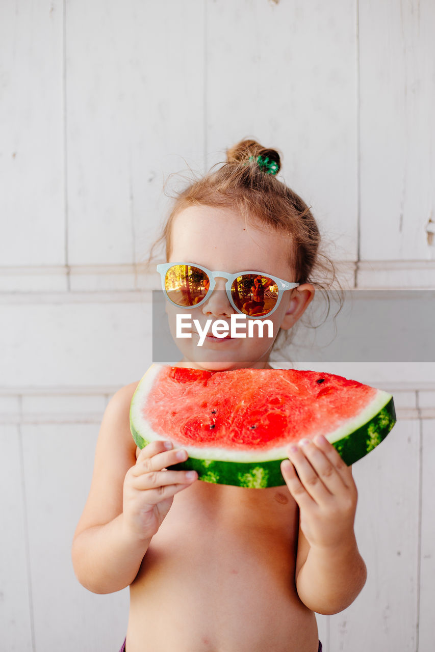 Portrait of shirtless girl eating watermelon against wall