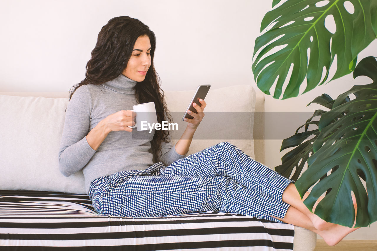 Beautiful  brunette woman browsing mobile phone and drinking coffee while resting on sofa at home.