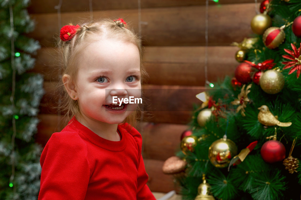 Portrait of a happy little girl in a red dress wooden background with a decorated christmas tree.