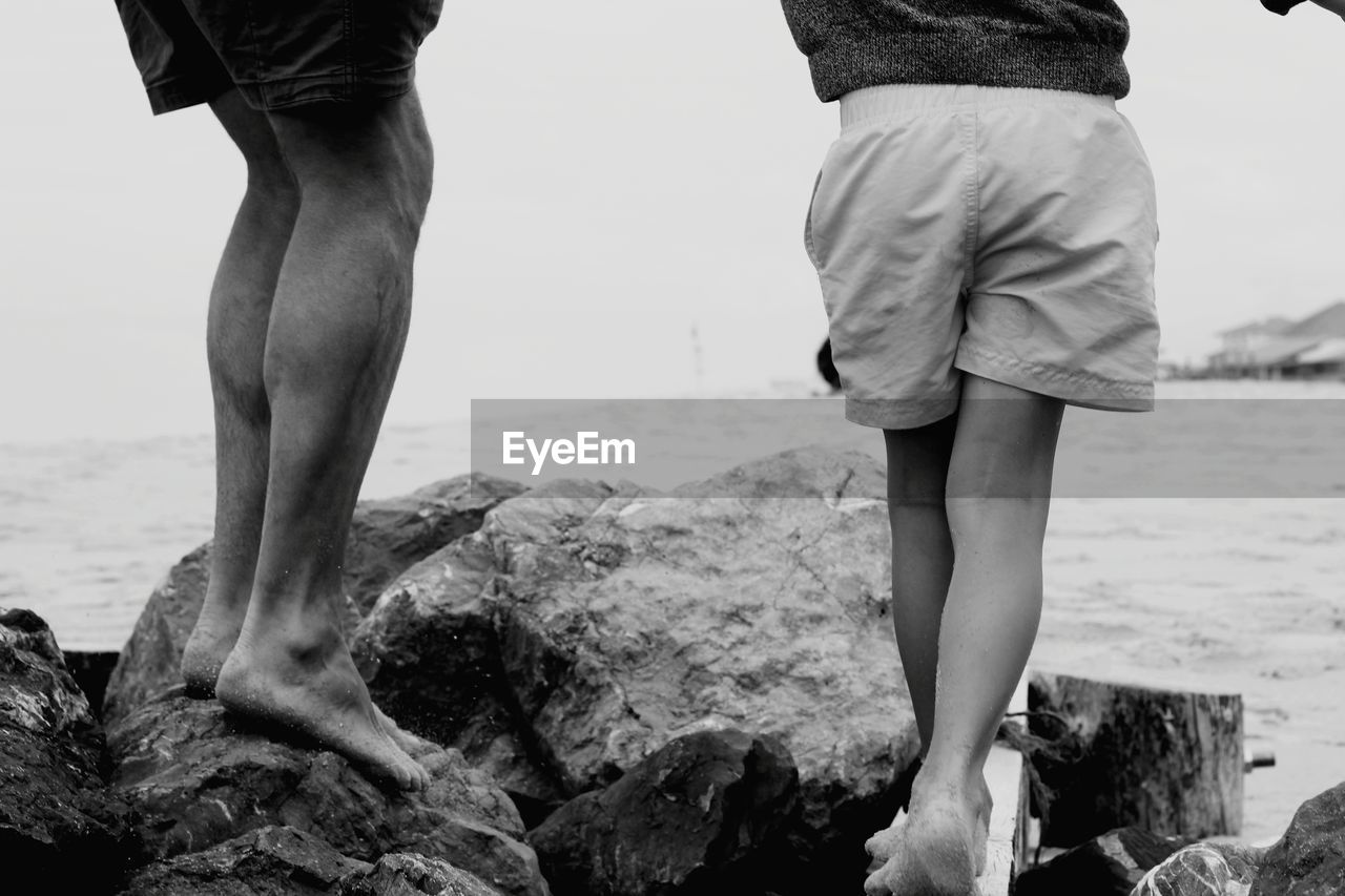 LOW SECTION OF MEN STANDING ON ROCK AT BEACH