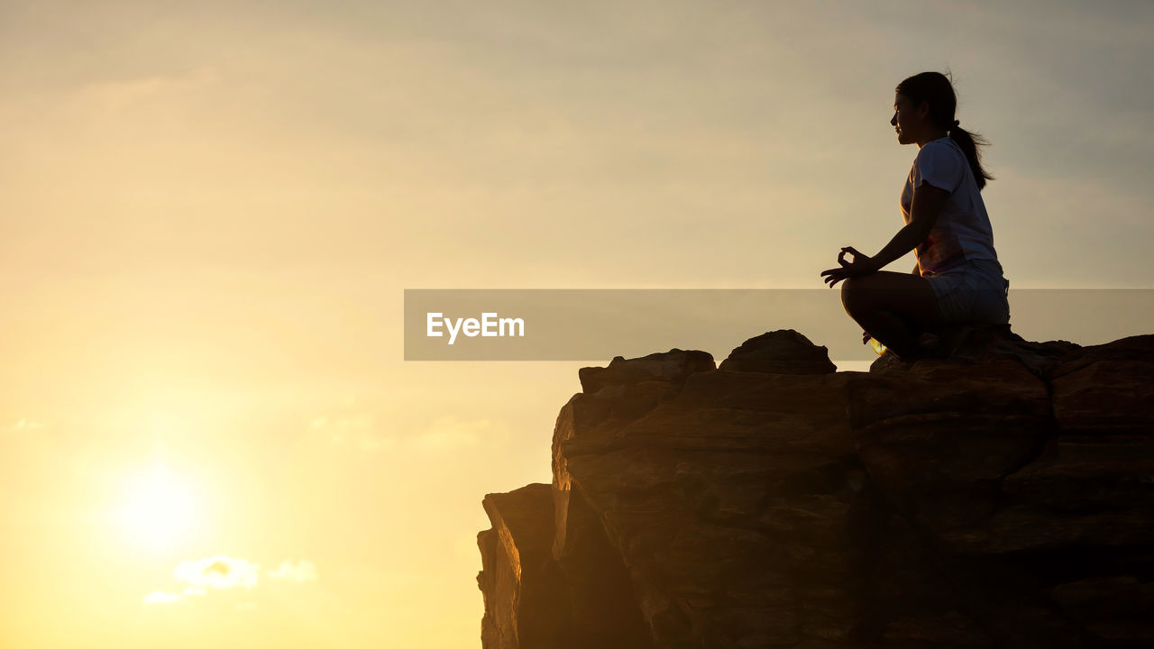 Low angle view of woman doing yoga on rock formation against sky during sunset