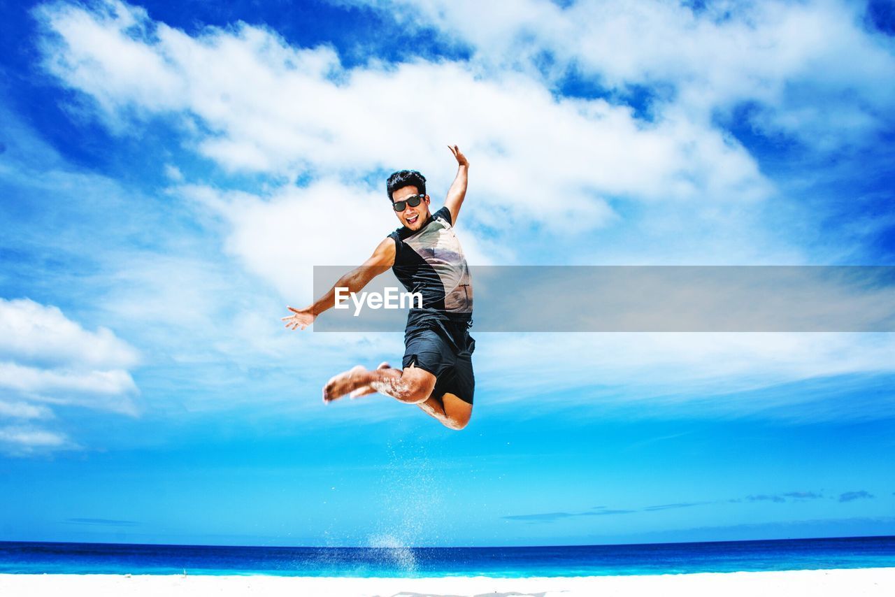 Happy young man jumping at beach against blue sky